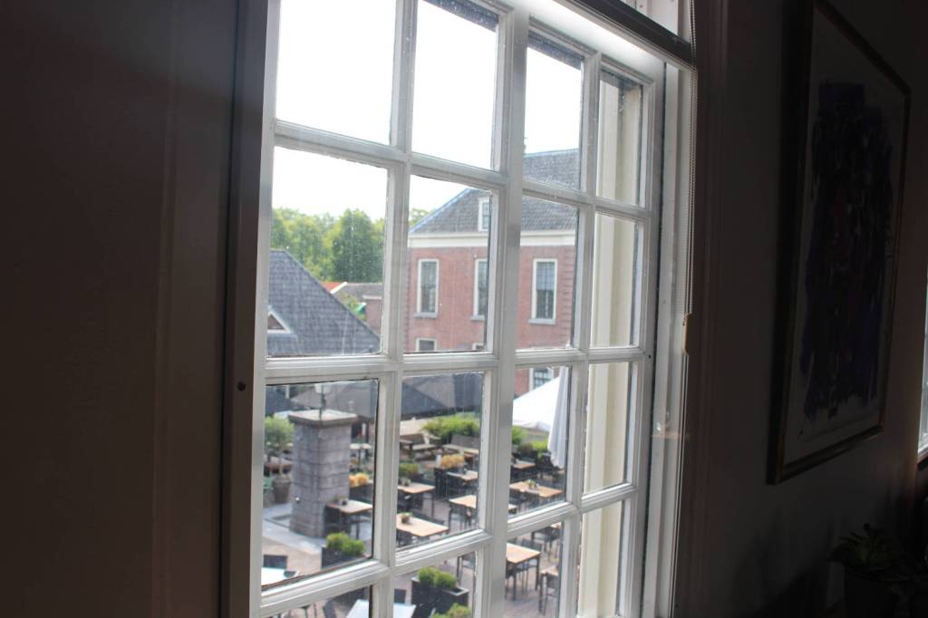 Uitzicht uit één van de ramen van de Kazerne. Behalve de tafeltjes zal het uitzicht niet veel veranderd zijn. De foto met de vlag is genomen bij de stenen column, zichtbaar in het raampje linksonder. Foto: marjolijn kok.
